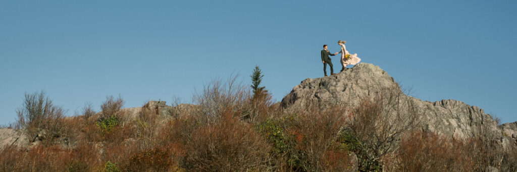 panorama of bride and groom climb on rocks in the Grayson Highlands of Virginia with blue sky